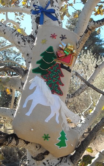 A Christmas stocking made of burlap with a white horse, a pine tree, and a sleigh with gifts