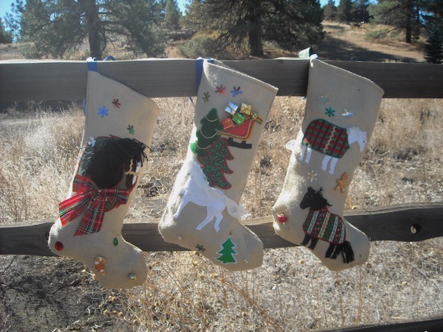 Three Christmas Stockings hanging on a fence. The stockings are burlap with applique. One has a brown horse head in a wreath, the next has a trotting white horse, a sleigh and a pine tree, the third has two horses one white, one black and they are wearing colorful Christmas blankets.